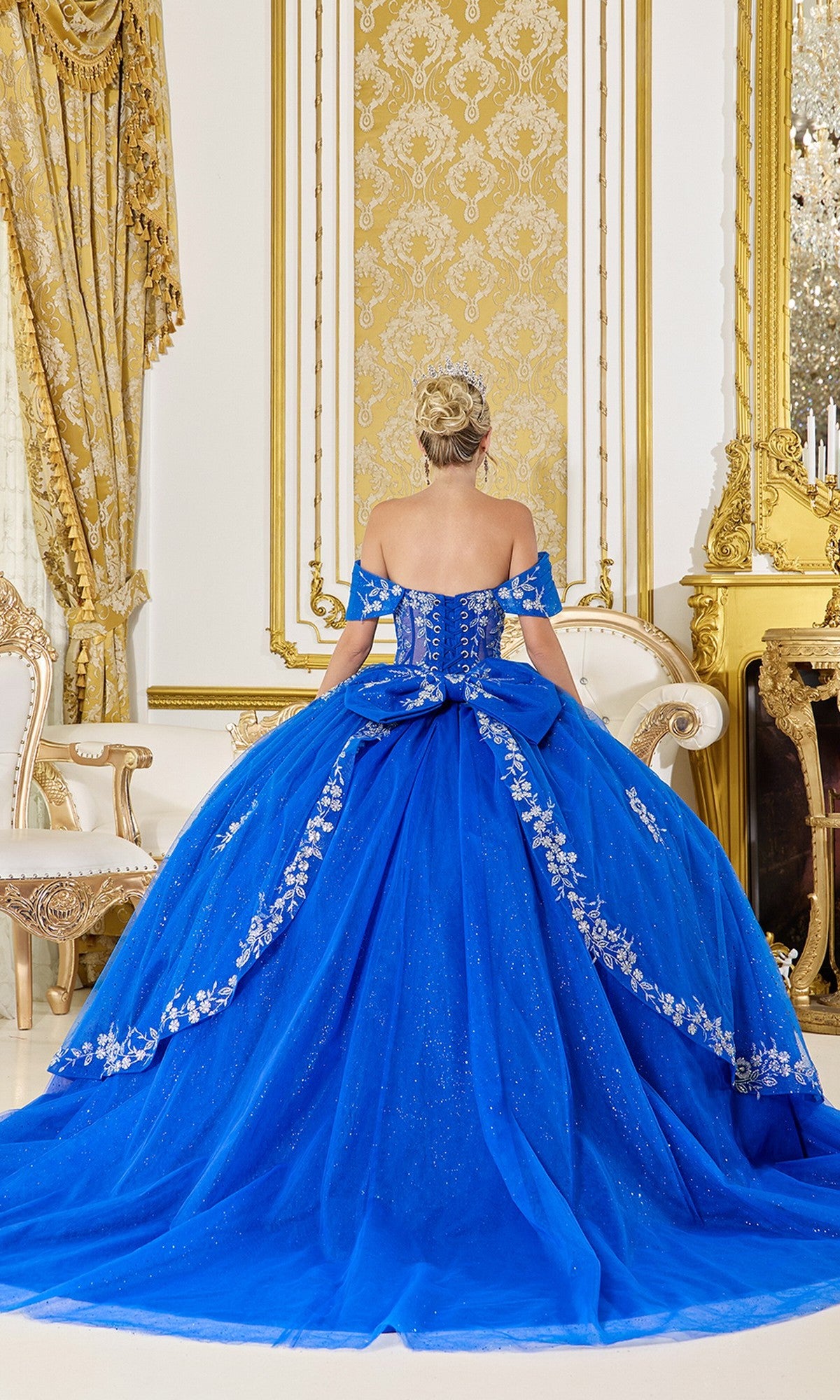 A woman in the Quinceanera Dress 5066 By Dancing Queen, featuring off-the-shoulder royal blue fabric with silver embroidery, stands facing away in an ornate room with gold accents.
